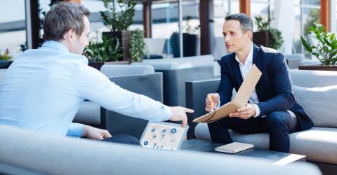 Man in pak wijst naar document in zijn hand aan man tegenover hem met een laptop voor zich